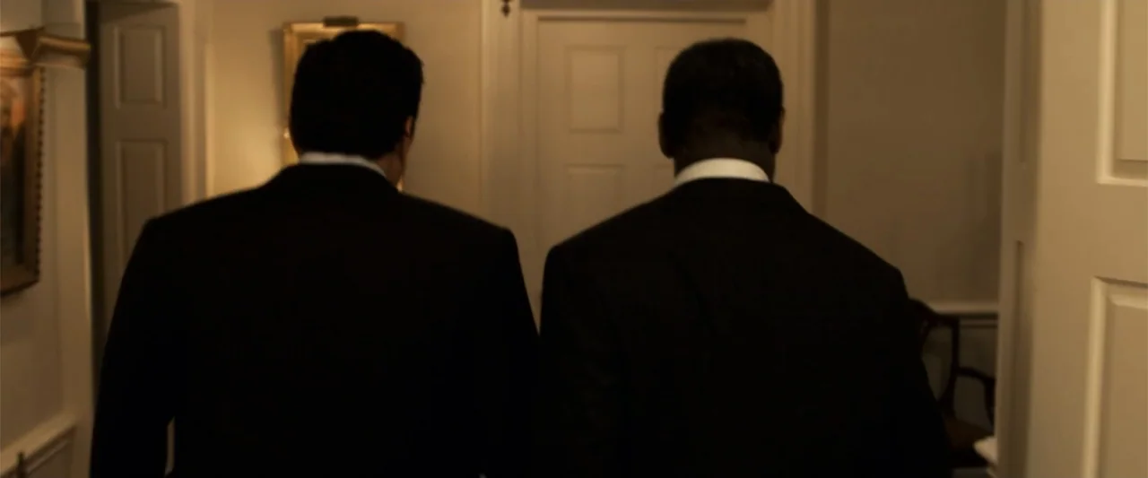 2012 (2009) movie still: medium — A dimly lit indoor hallway with white walls and a closed door ahead. Two men in dark suits walk away from the…
