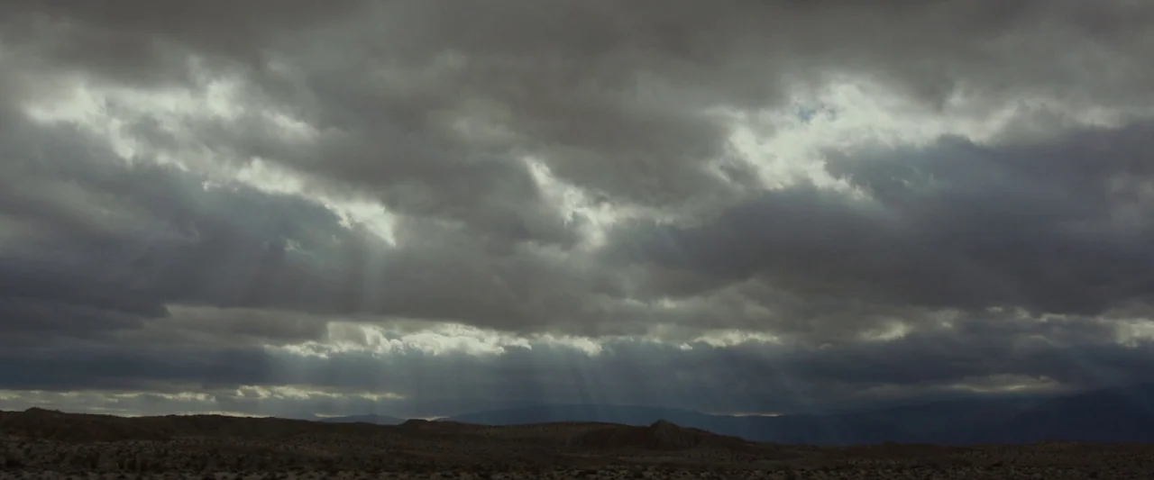 Last Days in the Desert (2015) movie still: establishing — An arid, rocky desert landscape sits beneath a vast expanse of dark, heavy clouds. Light…