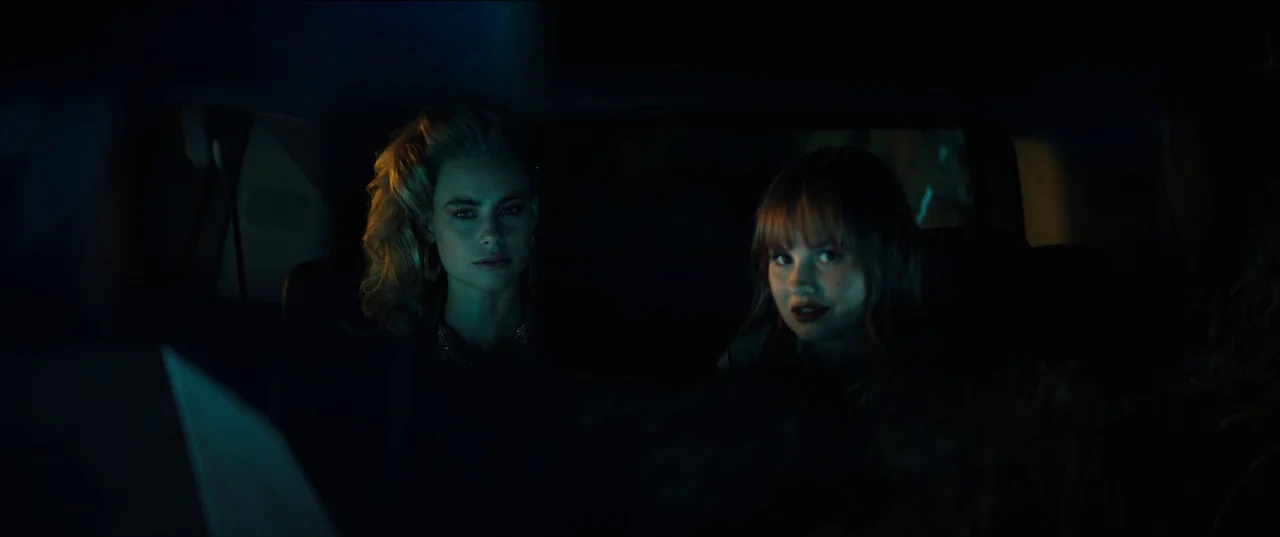 Night Teeth (2021) movie still: medium — The interior of a vehicle at night is illuminated by passing exterior light. Two women sit in the backseat…