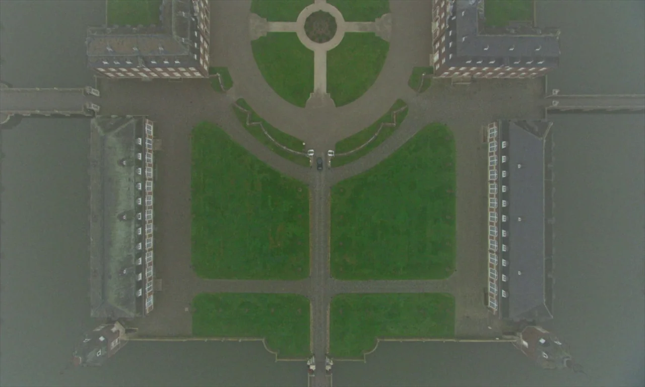 Spencer (2021) movie still: establishing — A symmetric castle complex surrounds a central courtyard with green lawns and stone paths. A single dark…