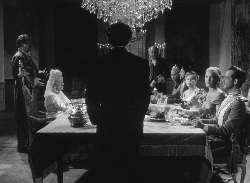 Smiles Of A Summer Night (1955) movie still: medium — A group of people sits at a long, formally set dining table in a manor. A man stands at the…
