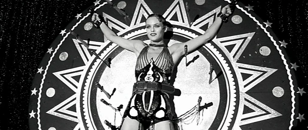 Girl on the Bridge (1999) movie still: medium — A woman stands strapped to a large, circular, star-adorned spinning target board. Multiple sharp…