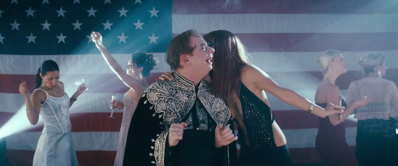 Southland Tales (2006) movie still: medium — A dance floor is decorated with a large American flag backdrop. A man in an ornate cape dances next to…