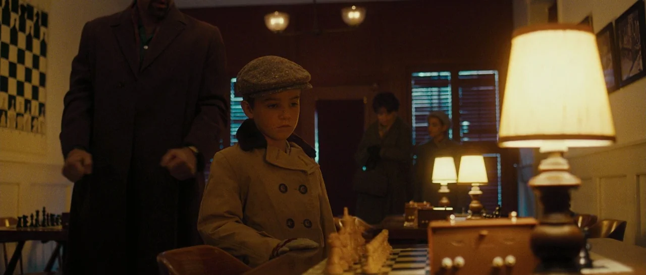 Pawn Sacrifice (2014) movie still: medium — A dark, wood-paneled room is dimly lit by small lamps placed on tables. A young boy wearing a cap stands…