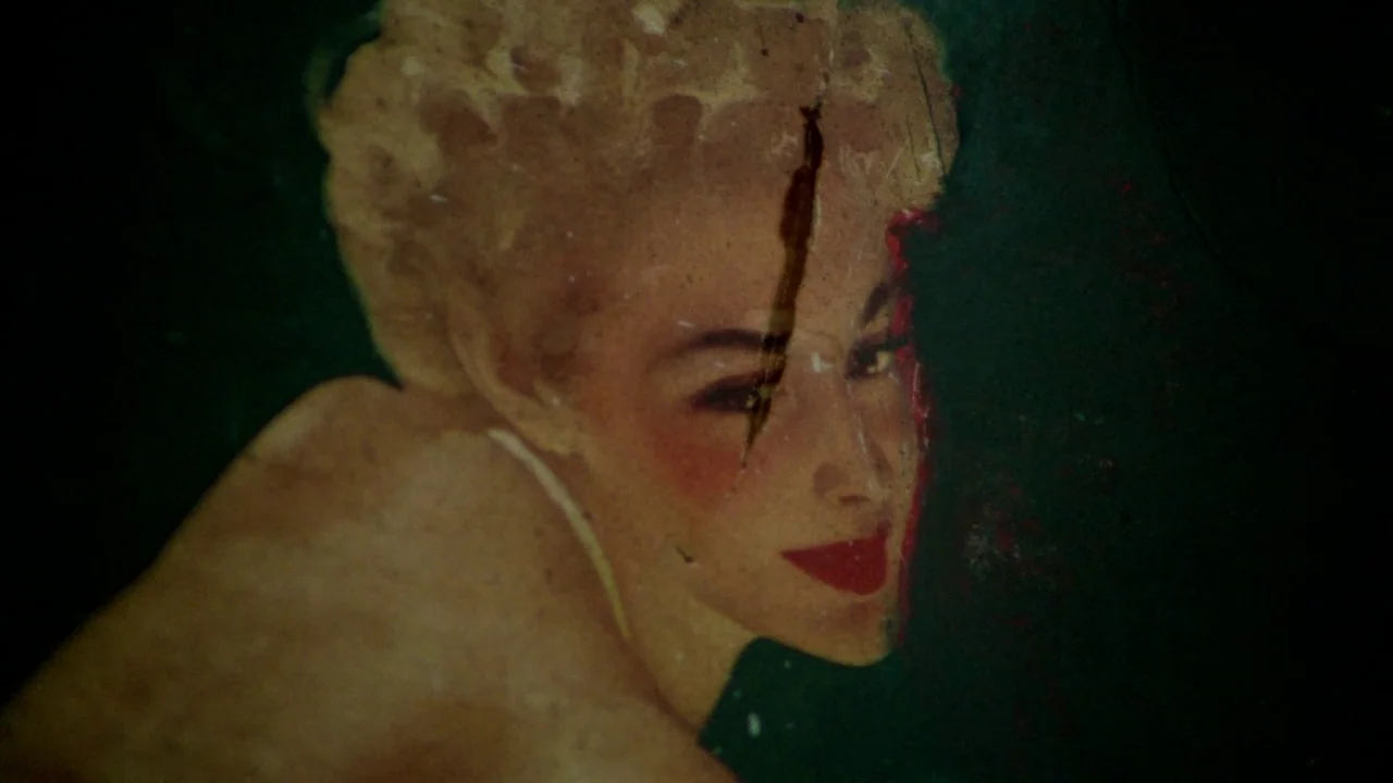 Sorcerer (1977) movie still: closeup — A worn, scratched vintage illustration of a woman is fixed to a dark surface. The image shows a blonde woman…