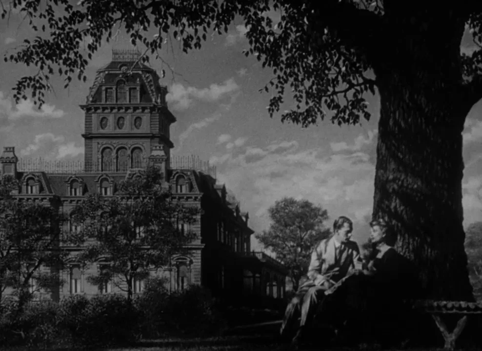 The Magnificent Ambersons (1942) movie still: wide — A man and woman sit on a bench under a large, leafy tree. In the background stands a sprawling…