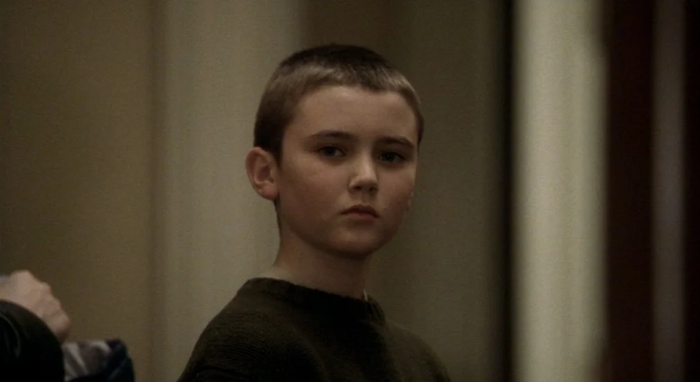Birth (2004) movie still: closeup — A neutral-toned indoor environment with plain, light-colored walls in the background. A young boy in a green…