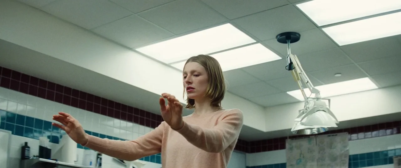 Kinds of Kindness (2024) movie still: medium — An examination room features white walls with dark red and teal accent tiling. A young woman holds…
