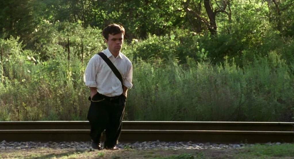 The Station Agent (2003) movie still: fullbody — A man walks alongside railroad tracks bordered by dense green brush and trees. He wears a dress…