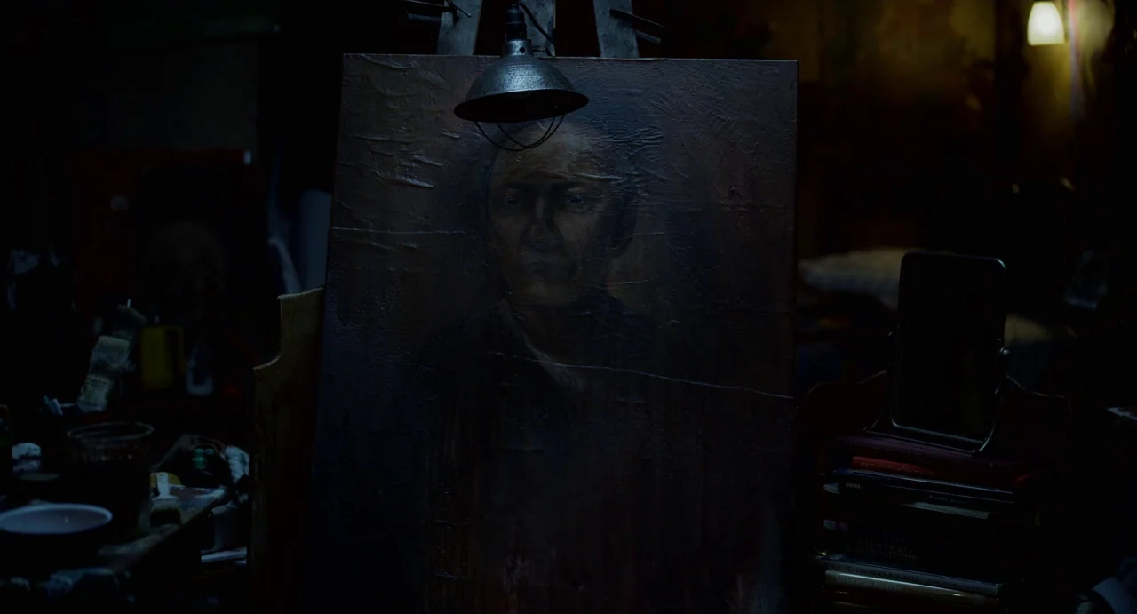Velvet Buzzsaw (2019) movie still: closeup — A dimly lit art studio contains a central easel supporting a dark oil painting. The portrait on the…