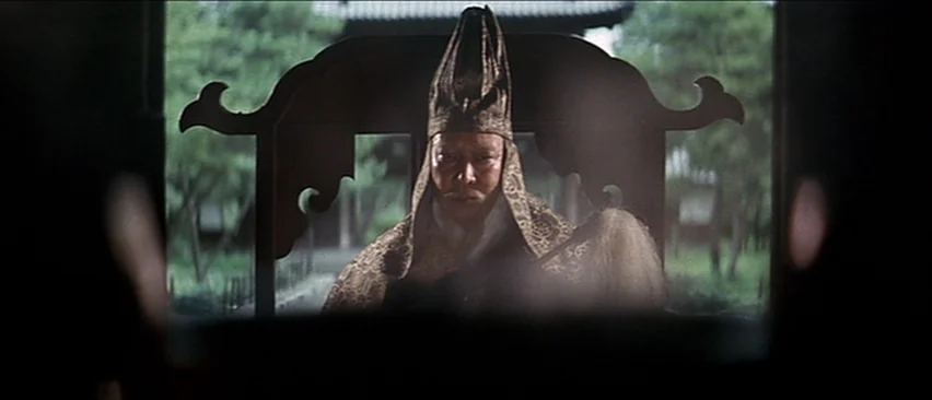 Lone Wolf and Cub: Baby Cart in the Land of Demons (1973) movie still: medium — An elderly figure in traditional attire sits in a wooden chair…