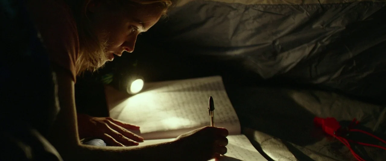 Wild (2014) movie still: closeup — A person lies inside a fabric structure at night with limited ambient light. The individual focuses on writing in…