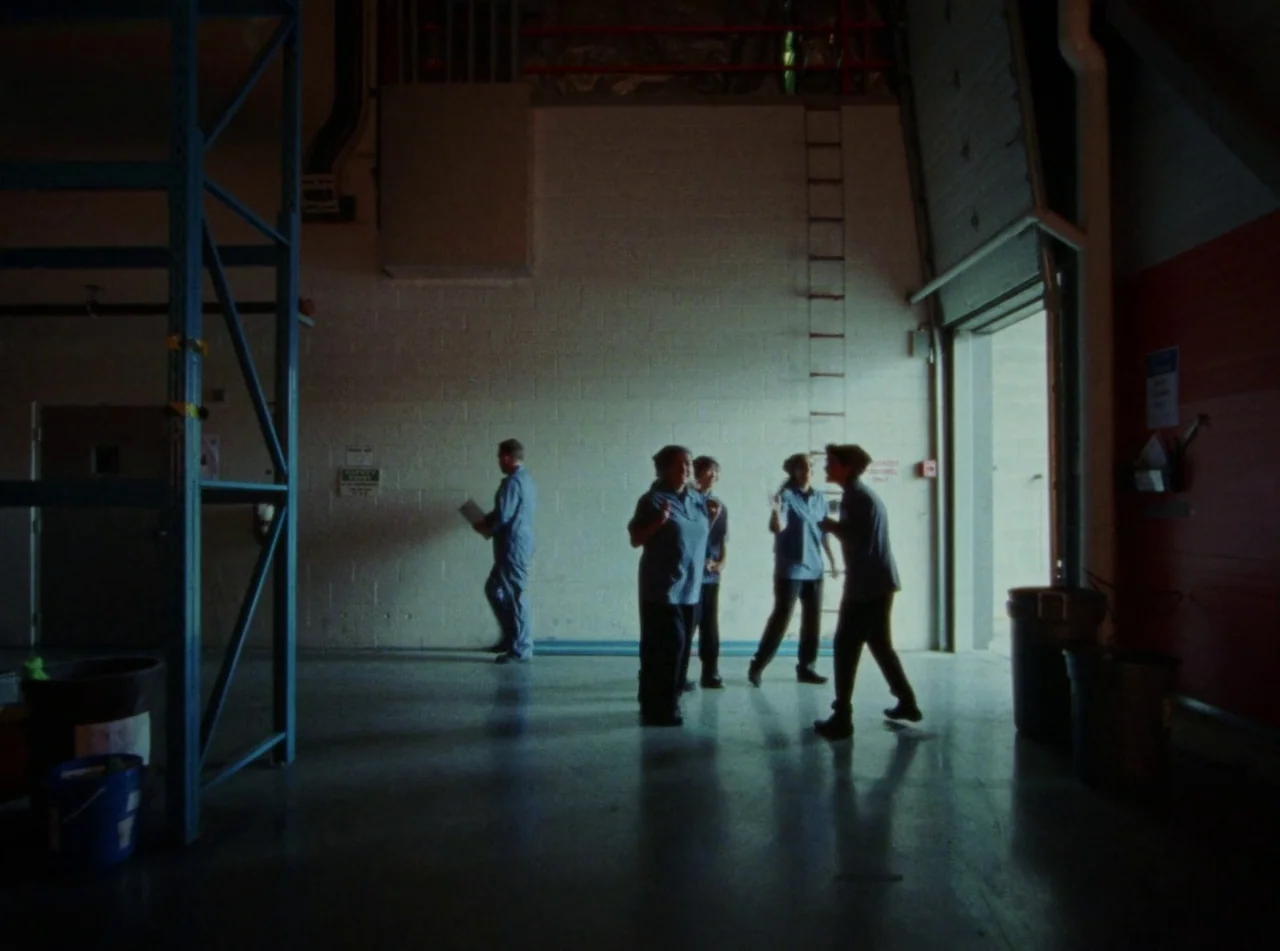Riceboy Sleeps (2022) movie still: wide — A high-ceilinged industrial storage room features white concrete block walls and metal shelving units. A…