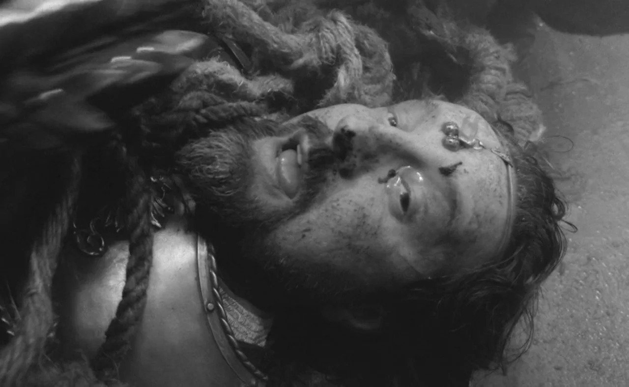 Hard To Be A God (2013) movie still: closeup — The face of a man in armor lies against a hard dusty surface. Thick rope is tangled around his…