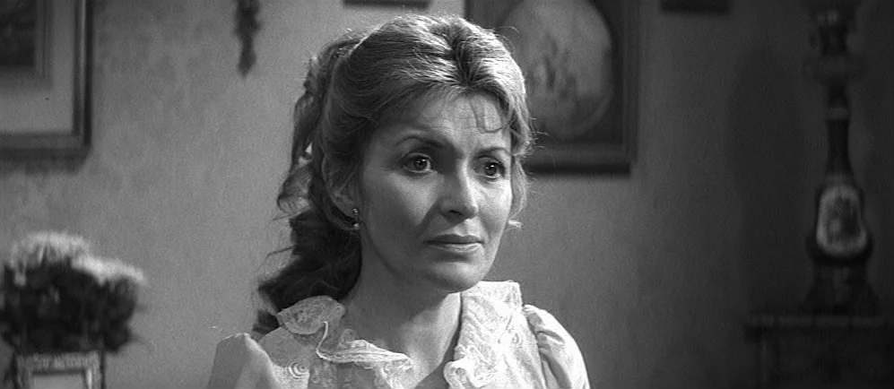 The Elephant Man (1980) movie still: closeup — A domestic space with framed wall decor and a decorative lamp base visible behind. A woman with wavy…