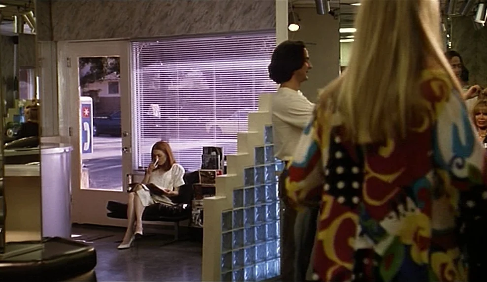 Safe movie still: medium — A 1990s hair salon features a glass block partition and a large window. A woman with auburn hair sits in a black chair…