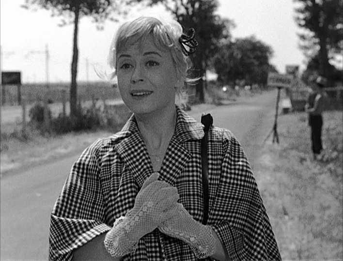 Nights of Cabiria (1957) movie still: medium — A woman stands on a country road bordered by trees and a distant sign. She holds her hands together…