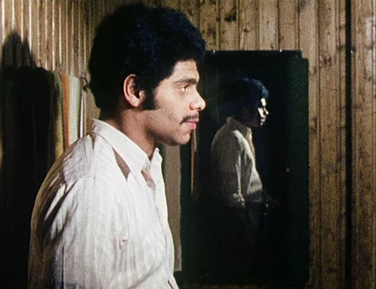 Rio Das Mortes (1971) movie still: medium — A small room with vertical wood paneled walls and a mirror. A young man stands in profile while looking…