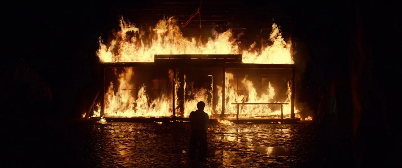 Never Grow Old (2019) movie still: wide — A wooden building is fully engulfed in flames in a dark outdoor setting at night. Two men stand in the…