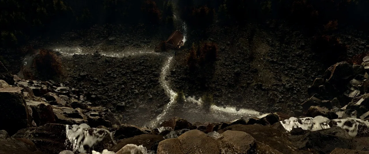 The House That Jack Built (2018) movie still: establishing — A stream carves a path through a rocky mountain slope dotted with pine trees. A small…