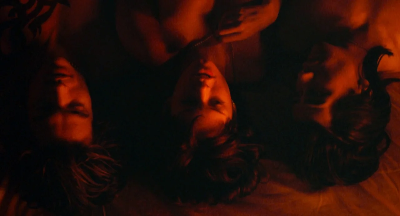 The Doom Generation (1995) movie still: medium — Three people lie head-to-head on a bed under low-light orange conditions. They are positioned in a…
