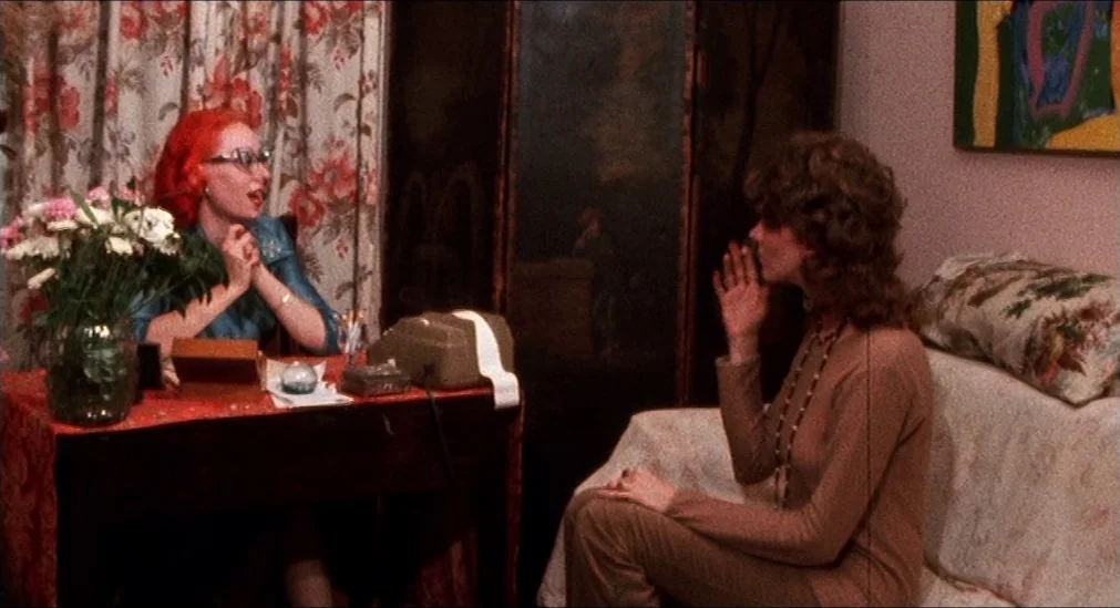 Pink Flamingos (1972) movie still: medium — A room contains a desk with a telephone and a small couch covered in pillows. Two women converse while…
