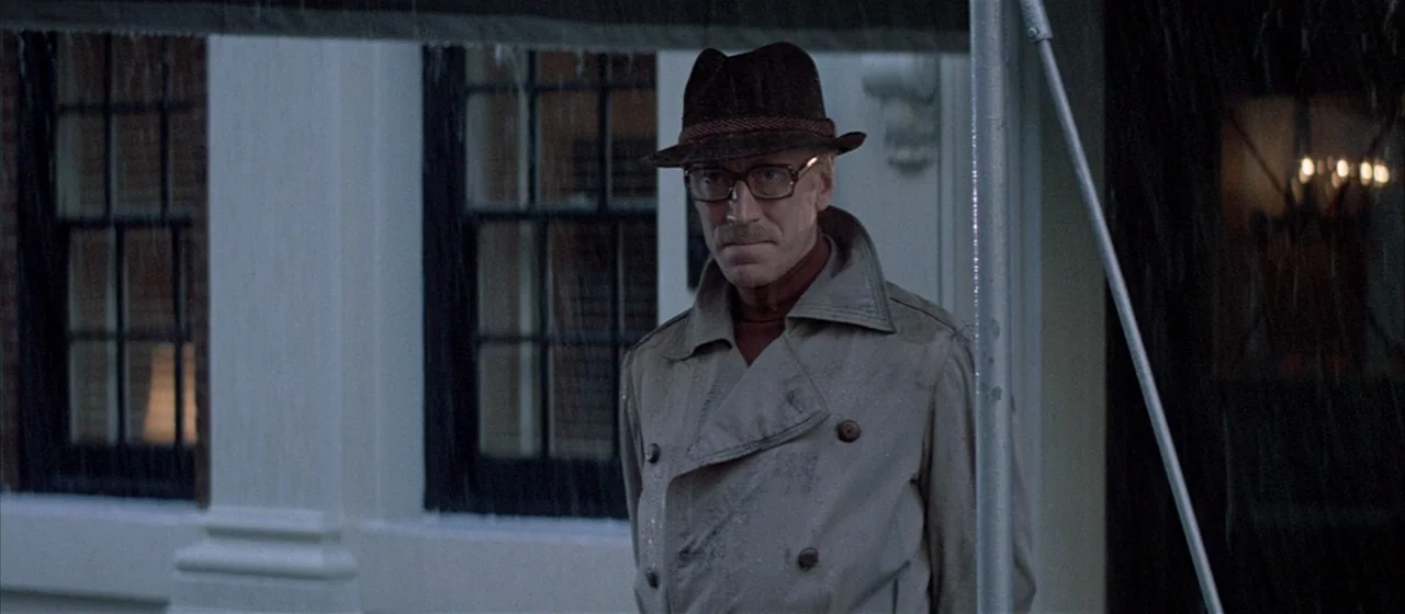 Three Days of the Condor (1975) movie still: medium — A man in a trench coat and hat stands before a white building facade. Rain falls steadily in…