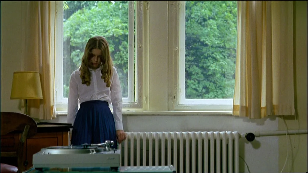 Chinese Roulette (1976) movie still: medium — A room with light-colored walls contains a window overlooking green foliage and trees. A girl stands…