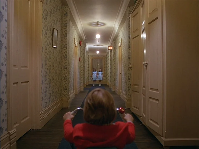 The Shining (1980) movie still: medium — A long, symmetrical hotel hallway features patterned wallpaper and multiple closed wooden doors. A child on…