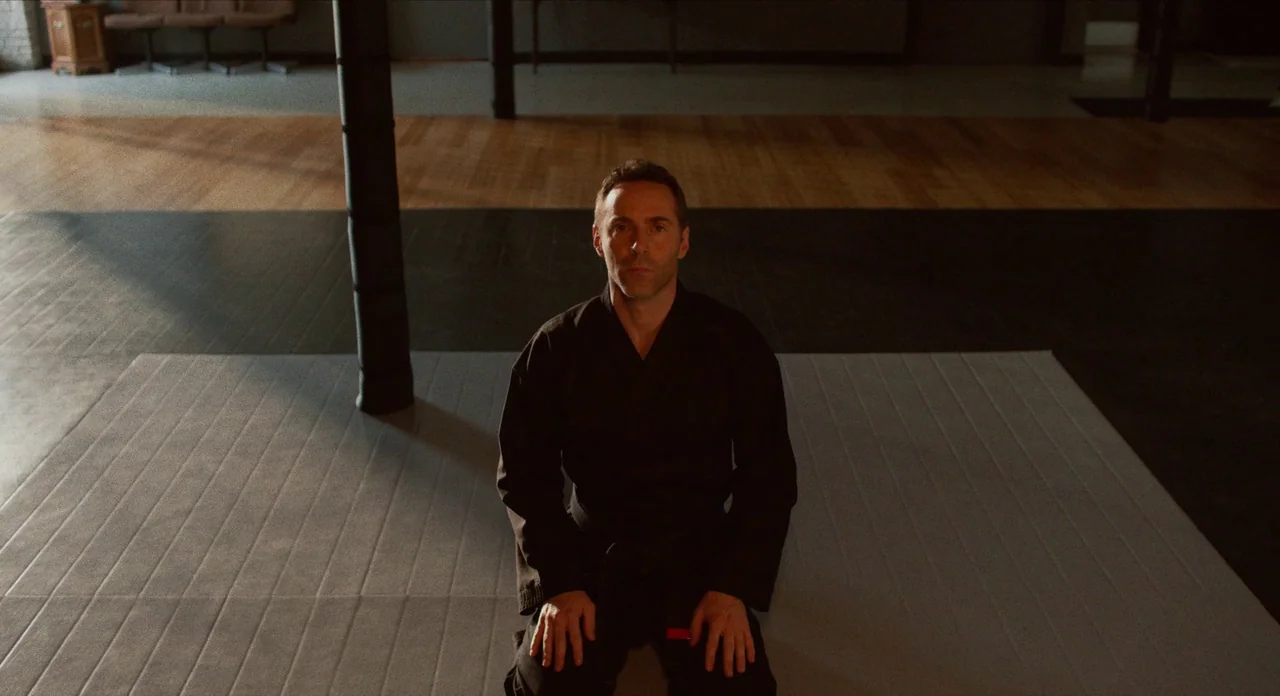 The Art of Self Defence (2019) movie still: medium — An indoor martial arts studio with a grey floor mat and dark wooden flooring. A man in a black…