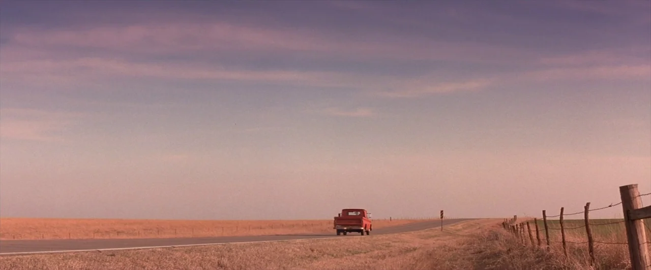 Mars Attacks! (1996) movie still: wide — A rural asphalt road cuts through flat, dry fields under a wide, pale sky. A lone red vintage pickup truck…