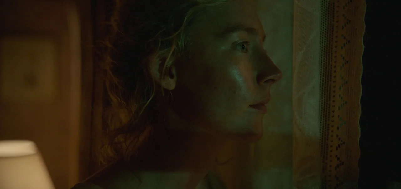 Foe (2023) movie still: closeup — A dimly lit room with green illumination reflecting off a woman's face. She stands by a curtained window gazing…