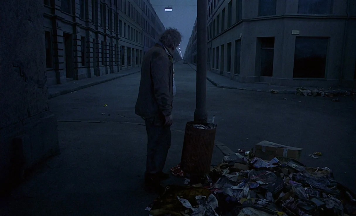 Songs from the Second Floor (2000) movie still: fullbody — A stark, empty urban street at night is lined with uniform, grey multi-story buildings.…