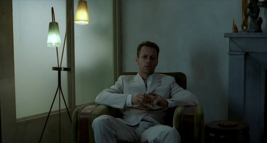 Anatomy of Hell (2004) movie still: medium — A sparsely furnished room features a green armchair and a tripod floor lamp. A man in a light blazer…