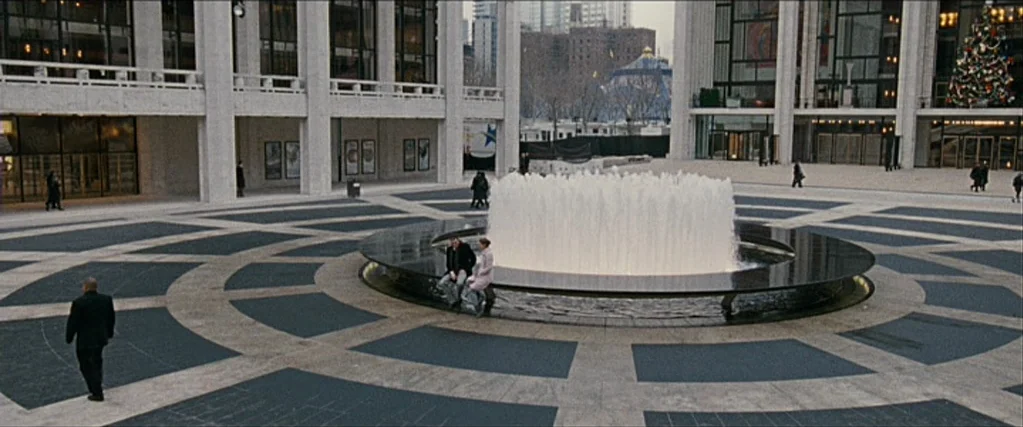 Black Swan (2010) movie still: wide — A wide concrete plaza surrounds a large circular water fountain with tall jets. Modern architectural buildings…
