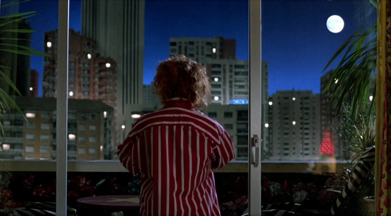 Kika (1993) movie still: medium — An interior space features a large glass sliding door looking out onto an urban skyline. A woman wearing a striped…