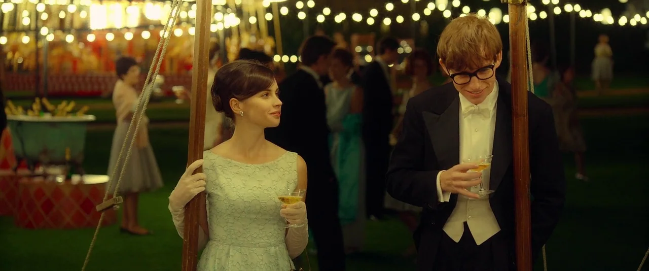 The Theory of Everything (2014) movie still: medium — A well-lit evening party takes place on a manicured lawn under draped string lights. A young…