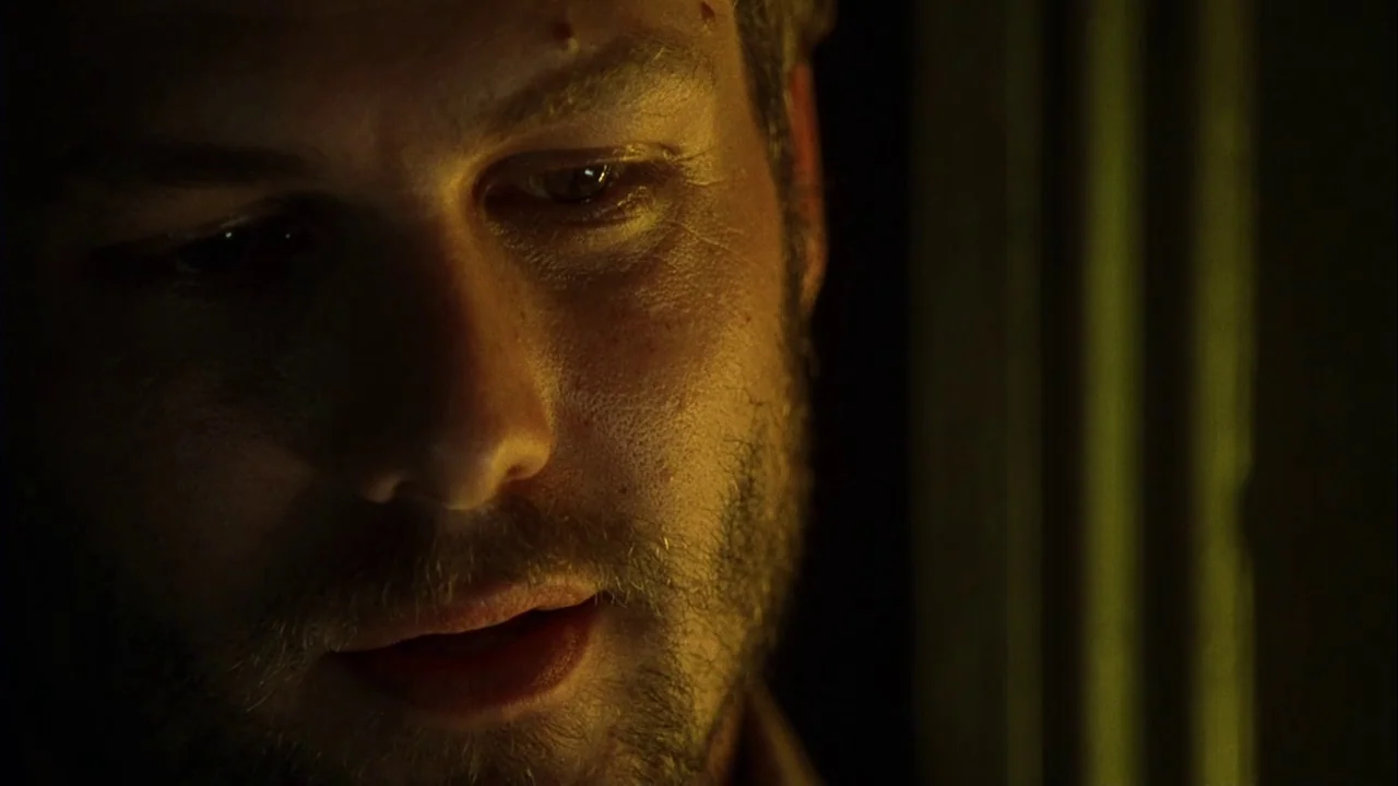 Love Song For Bobby Long (2004) movie still: closeup — A man is shown in a dark room with minimal architectural details visible. He looks down while…