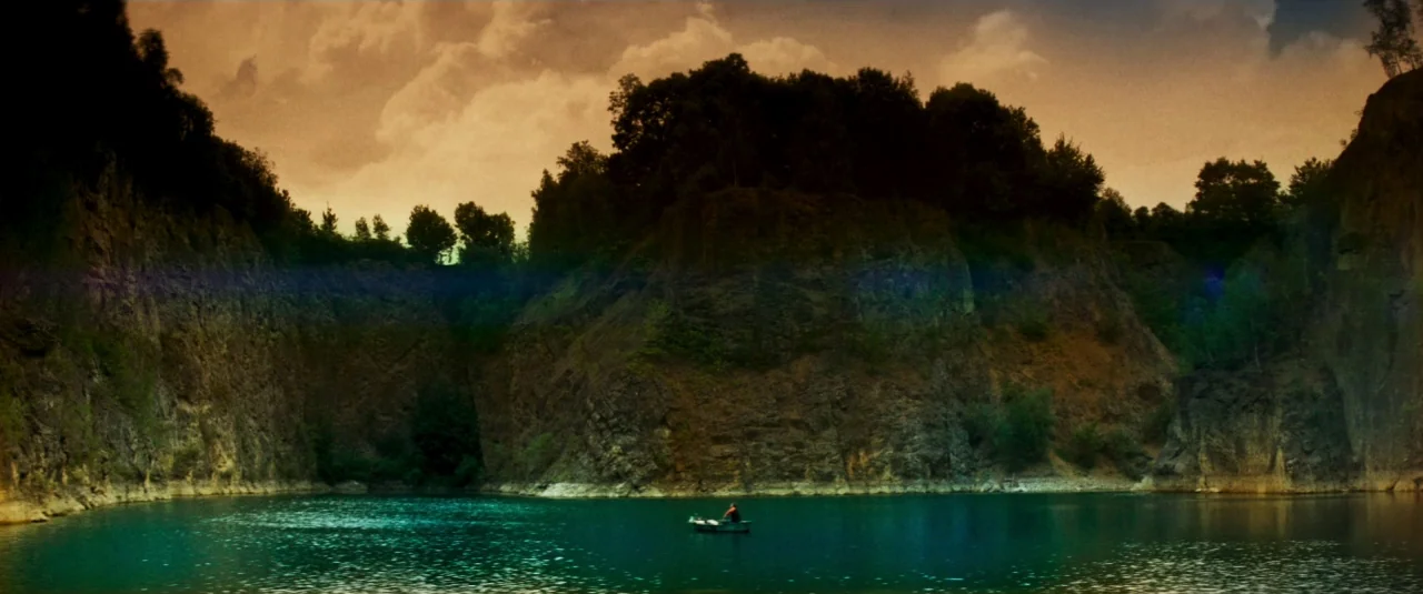 Mandy (2018) movie still: wide — A deep quarry lake is surrounded by steep, craggy rock walls covered in trees. A single small boat with a person…
