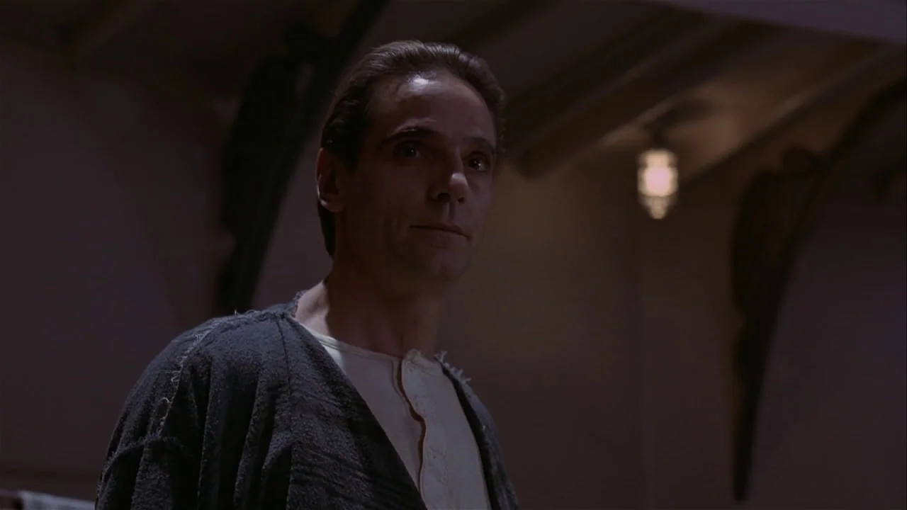 M. Butterfly (1993) movie still: medium — A dimly lit interior space with shadows obscuring most of the surrounding area. A man in a dark cardigan…