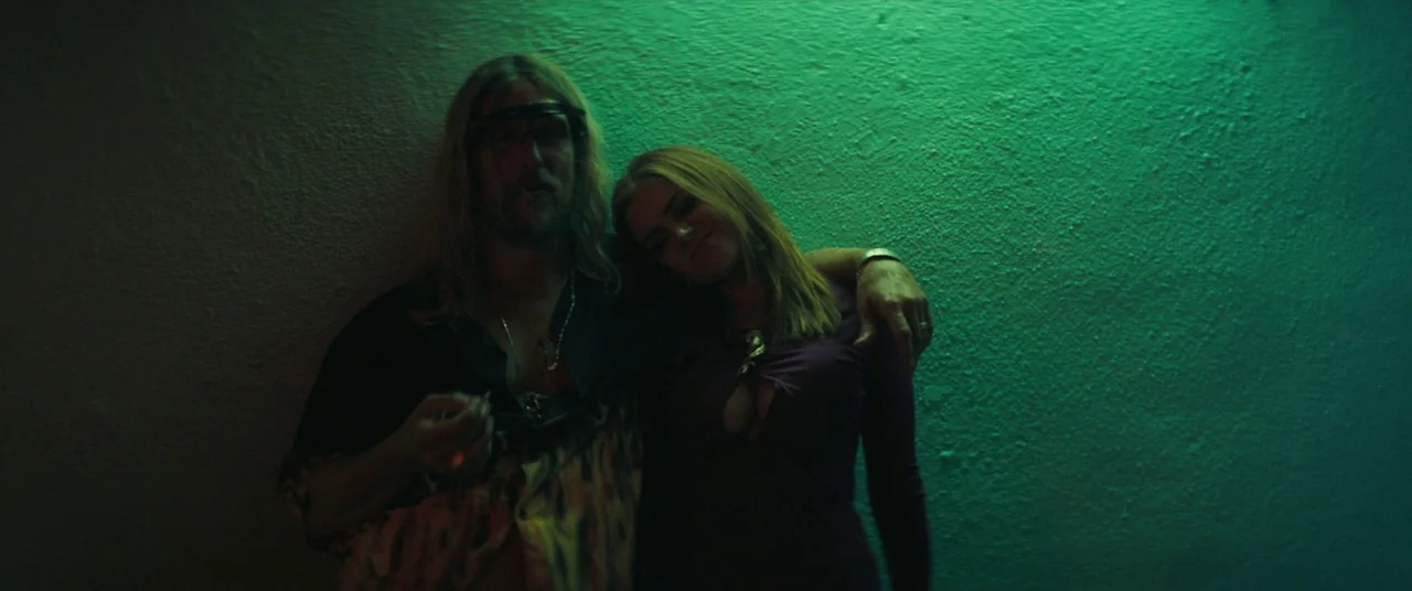 The Beach Bum (2019) movie still: medium — A dimly lit indoor space features a textured wall washed in vibrant green light. A man and a woman lean…