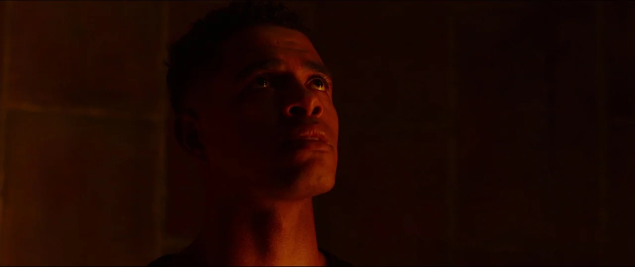 Jigsaw (2017) movie still: closeup — A man stands in a dark interior space with shadowed walls. He looks upward with a neutral expression while soft…