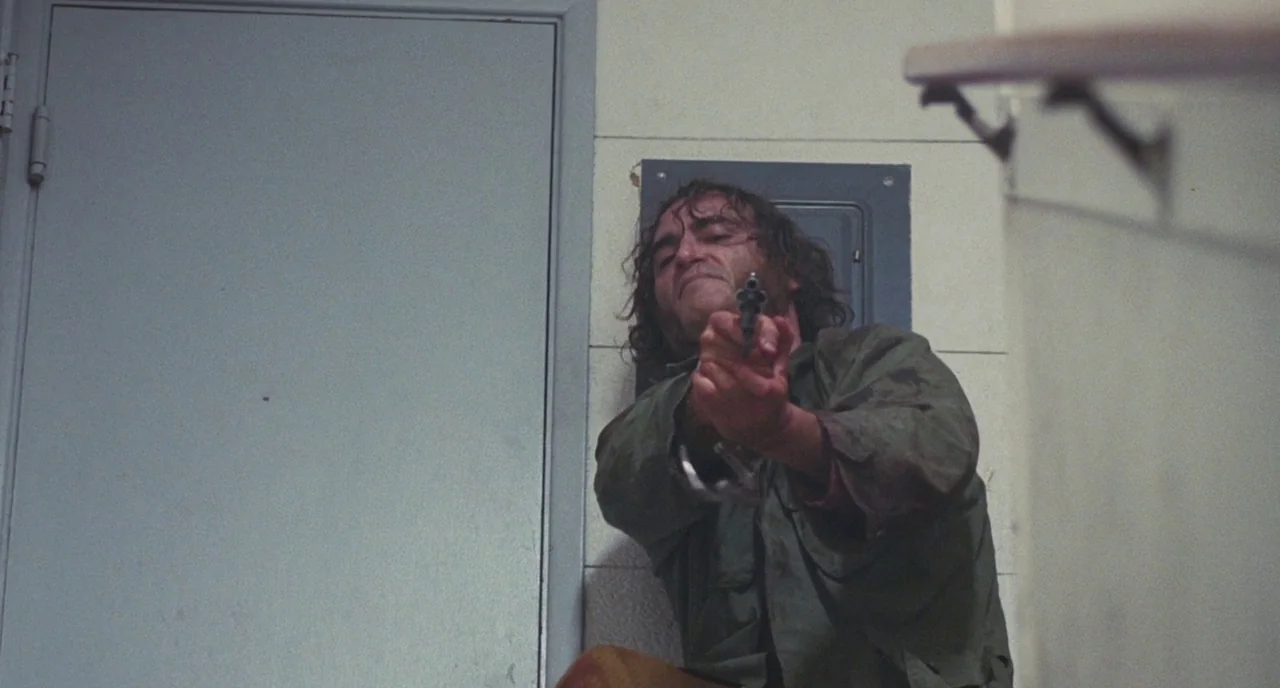 Inherent Vice (2014) movie still: medium — A stark stairwell with off-white tiled walls and a metal maintenance door. A disheveled man crouches on…