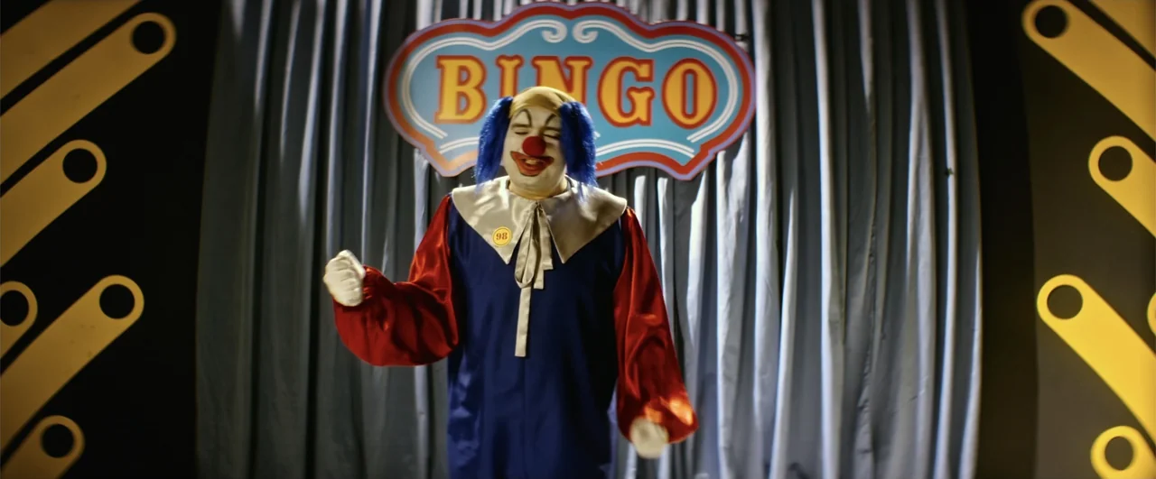 Bingo: The King of the Mornings (2017) movie still: medium — A clown in a costume stands in front of a blue curtain with a BINGO sign. The clown…