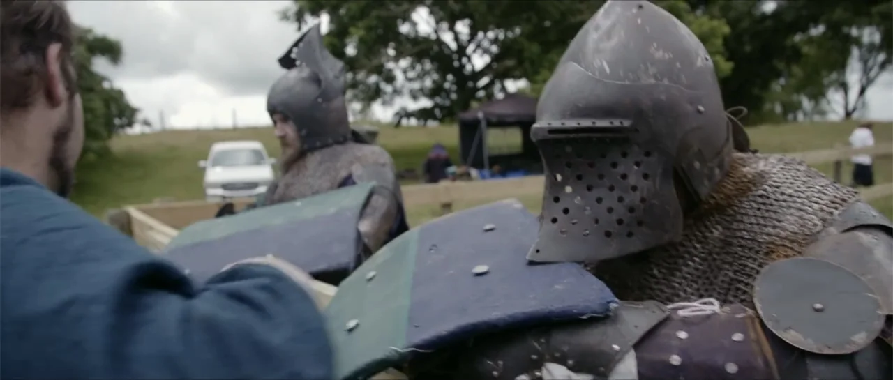 Bludgeon (2018) movie still: medium — A grassy outdoor training area with a wooden fence and a parked vehicle. Two armored individuals wearing…