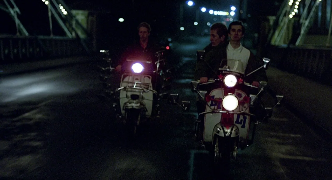 Quadrophenia (1979) movie still: medium — Three young men ride two vintage motor scooters across a dark, paved bridge at night. The scooters are…