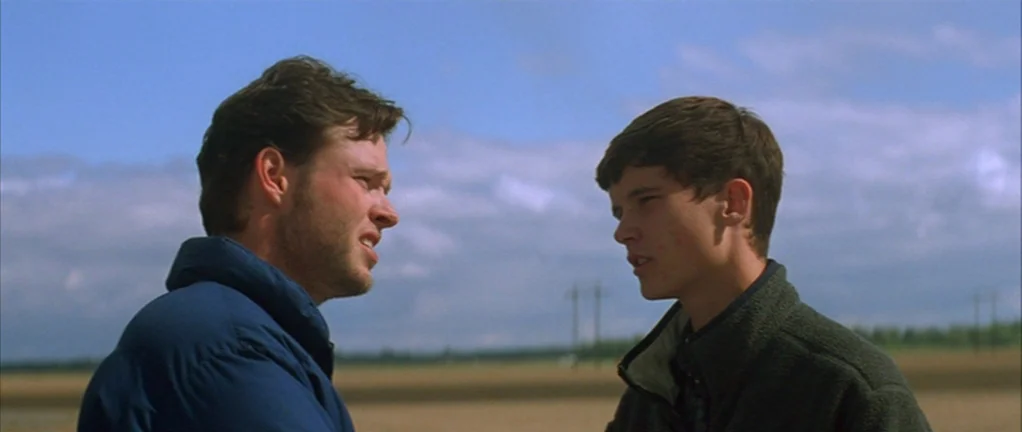 Shotgun Stories (2007) movie still: medium — An open rural field under a bright, partly cloudy blue sky. Two men in fleece jackets stand facing each…