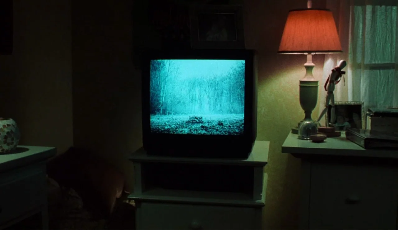 The Ring (2002) movie still: medium — A dimly lit room contains a CRT television sitting on a light-colored wooden cabinet. The television screen…