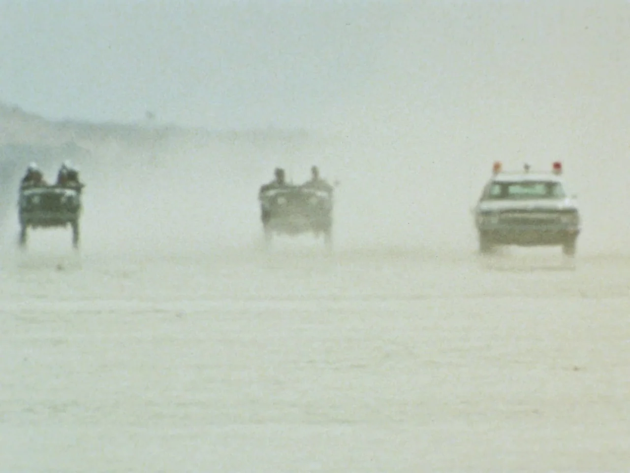 Punishment Park (1971) movie still: wide — A vast, flat expanse of white salt dust fills the frame under hazy light. Three vehicles move in parallel…