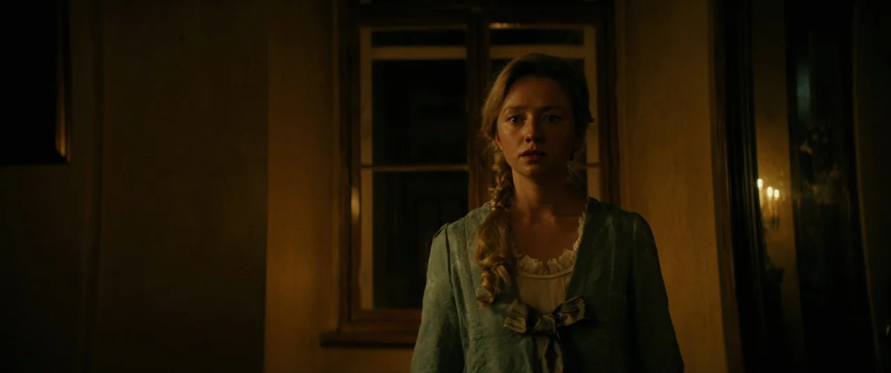 The Promised Land (2023) movie still: medium — A dimly lit room at night features a window centered behind a young woman. The subject stands facing…